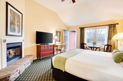A cozy hotel room with a fireplace, TV, desk, window, and a large bed with green pillows.