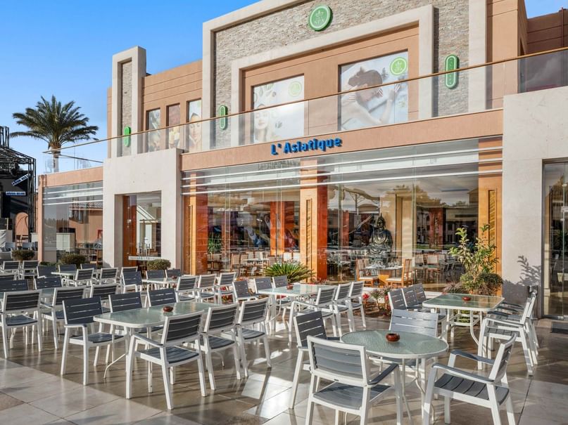 Außenbereich eines modernen Gebäudes mit Tischen und Stühlen, umgeben von Pflanzen und einer Glasfassade.