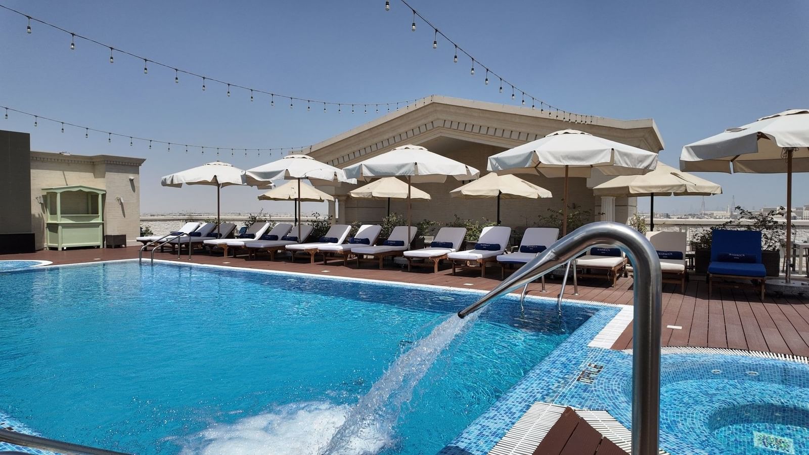 White loungers by a sparkling blue pool under patio umbrellas and hanging lights at Warwick Doha Hotel