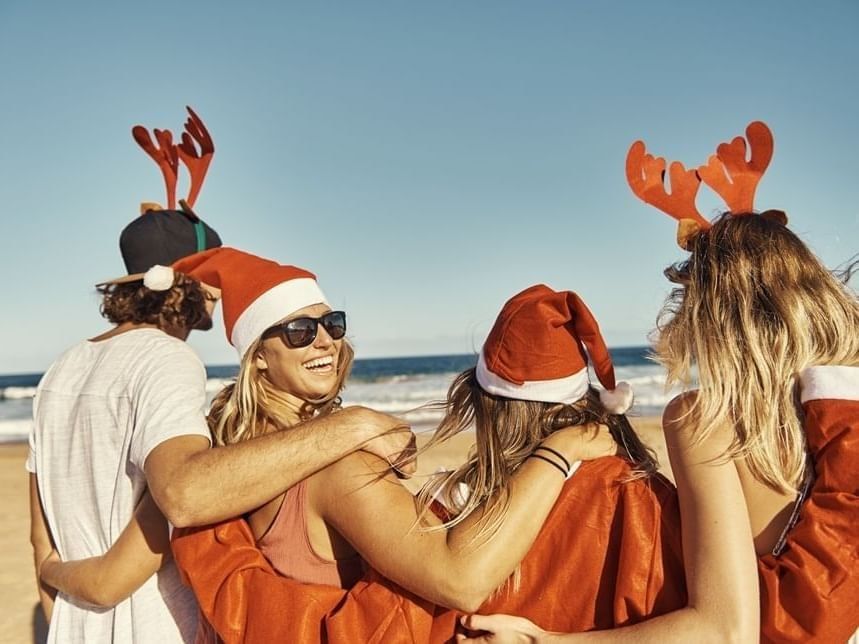 Four friends wearing festive Santa hats and reindeer antlers embrace on a sunny beach with ocean waves in the background.