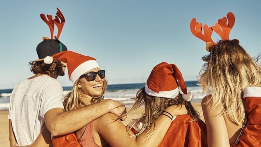 Four friends wearing festive Santa hats and reindeer antlers embrace on a sunny beach with ocean waves in the background.