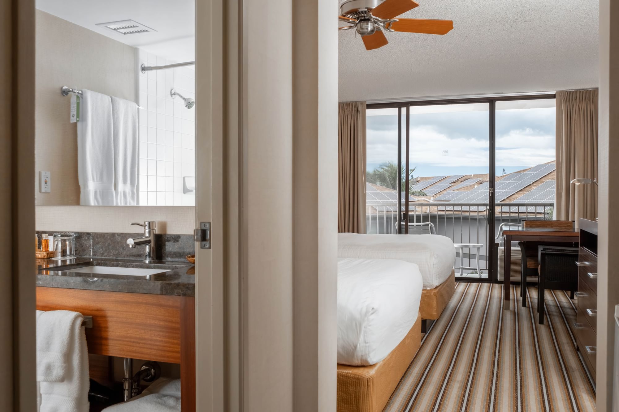 A hotel room with two beds, a desk, and a bathroom, with a view of the outdoors through a sliding glass door.
