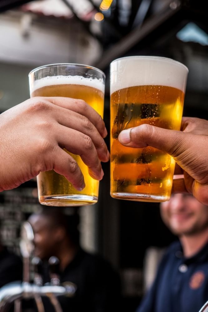 A closeup of two hands toasting with full glasses of pale beer. 