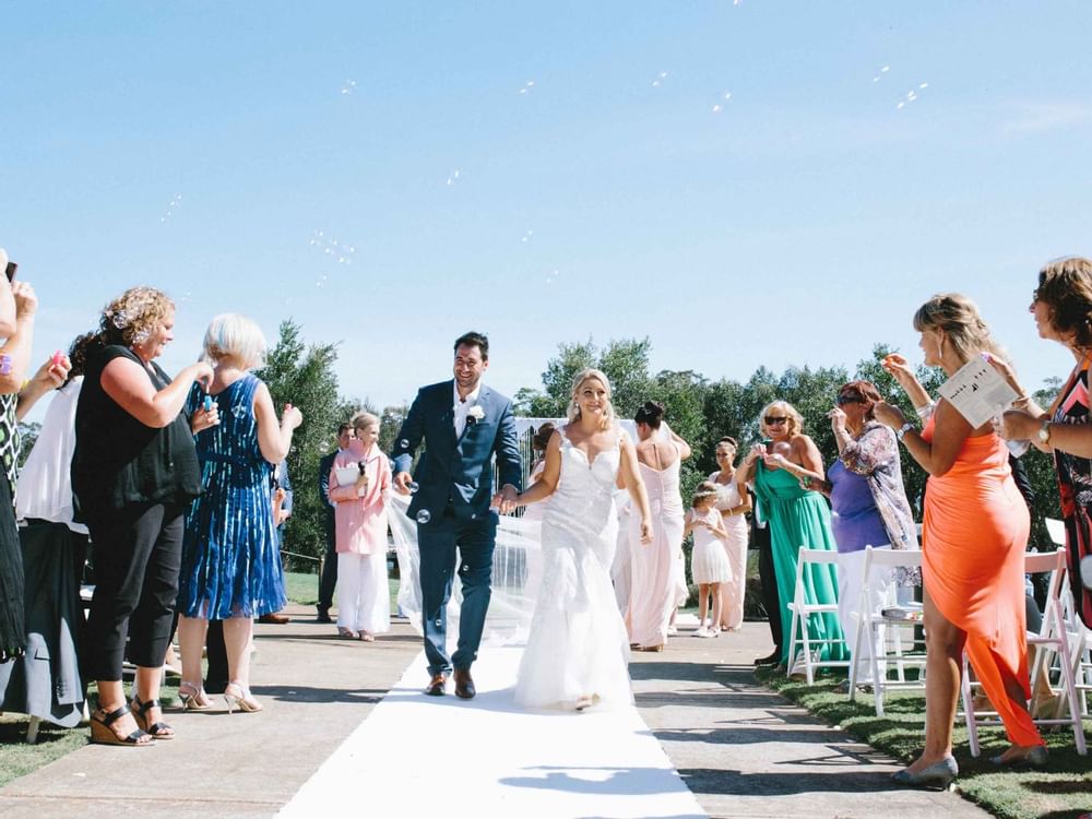 Couple walking down the aisle with guests blowing bubbles at an outdoor wedding ceremony in Mercure Kooindah Waters