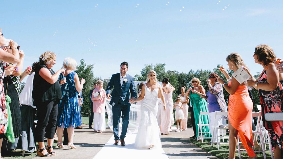 Couple walking down the aisle with guests blowing bubbles at an outdoor wedding ceremony in Mercure Kooindah Waters