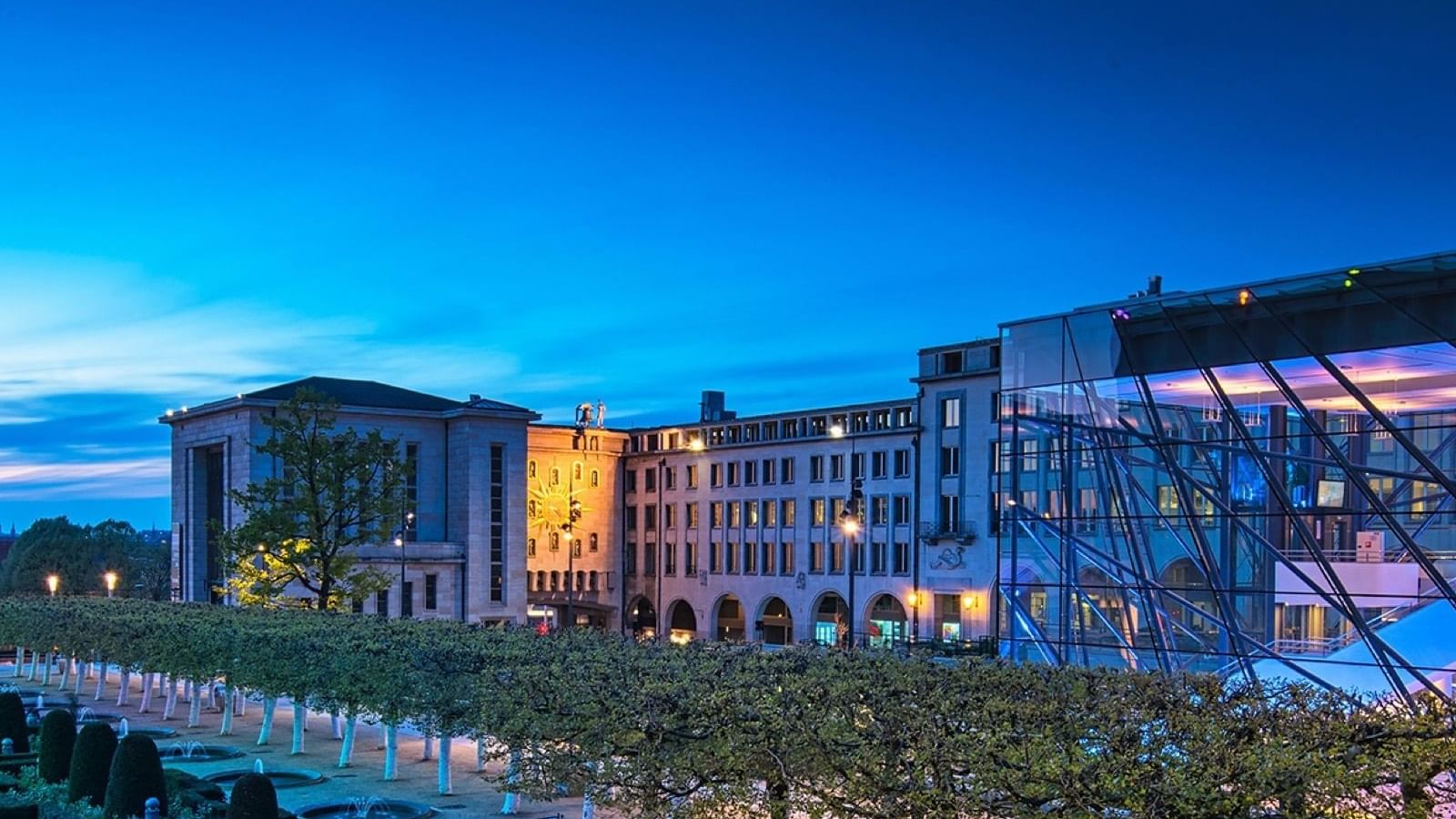 Modern plaza with manicured trees by a glass building and a gold clock at Hotel Barsey by Warwick