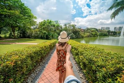 Woman in straw hat walks hand-in-hand by a serene lake on a sunny day at The Saujana Hotel Kuala Lumpur