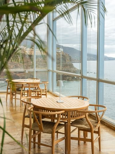 Wooden tables and chairs with lamps, large glass windows with view of the ocean and cliffs.