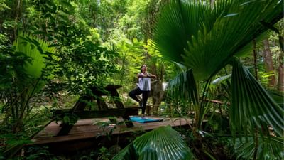Woman in tree pose on a yoga mat over a wooden deck surrounded by lush green tropical trees at Sugar Cane Club Hotel & Spa