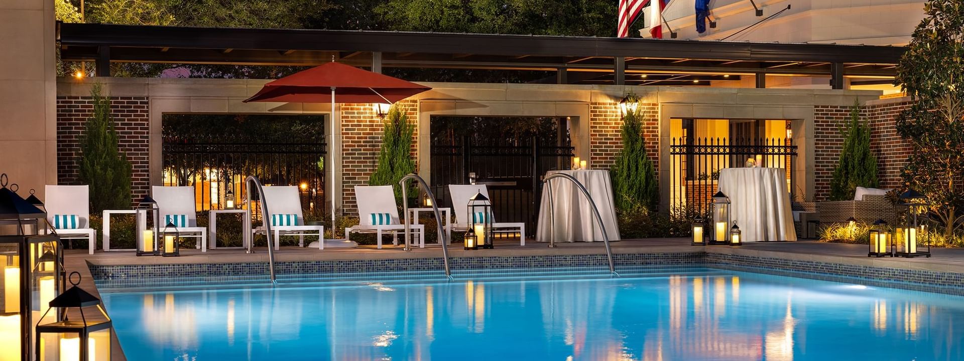 Illuminated pool Side by lounge chairs under a red umbrella near a brick building at Warwick Melrose Dallas