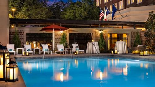 Illuminated pool Side by lounge chairs under a red umbrella near a brick building at Warwick Melrose Dallas