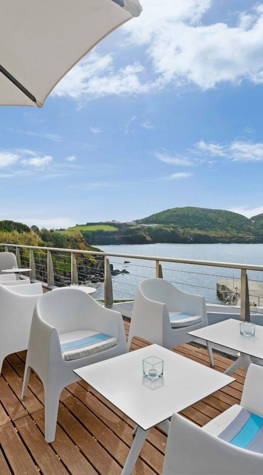 Terrace with white chairs and tables under large umbrellas on a wooden deck overlooking the bay at Hotel do Caracol