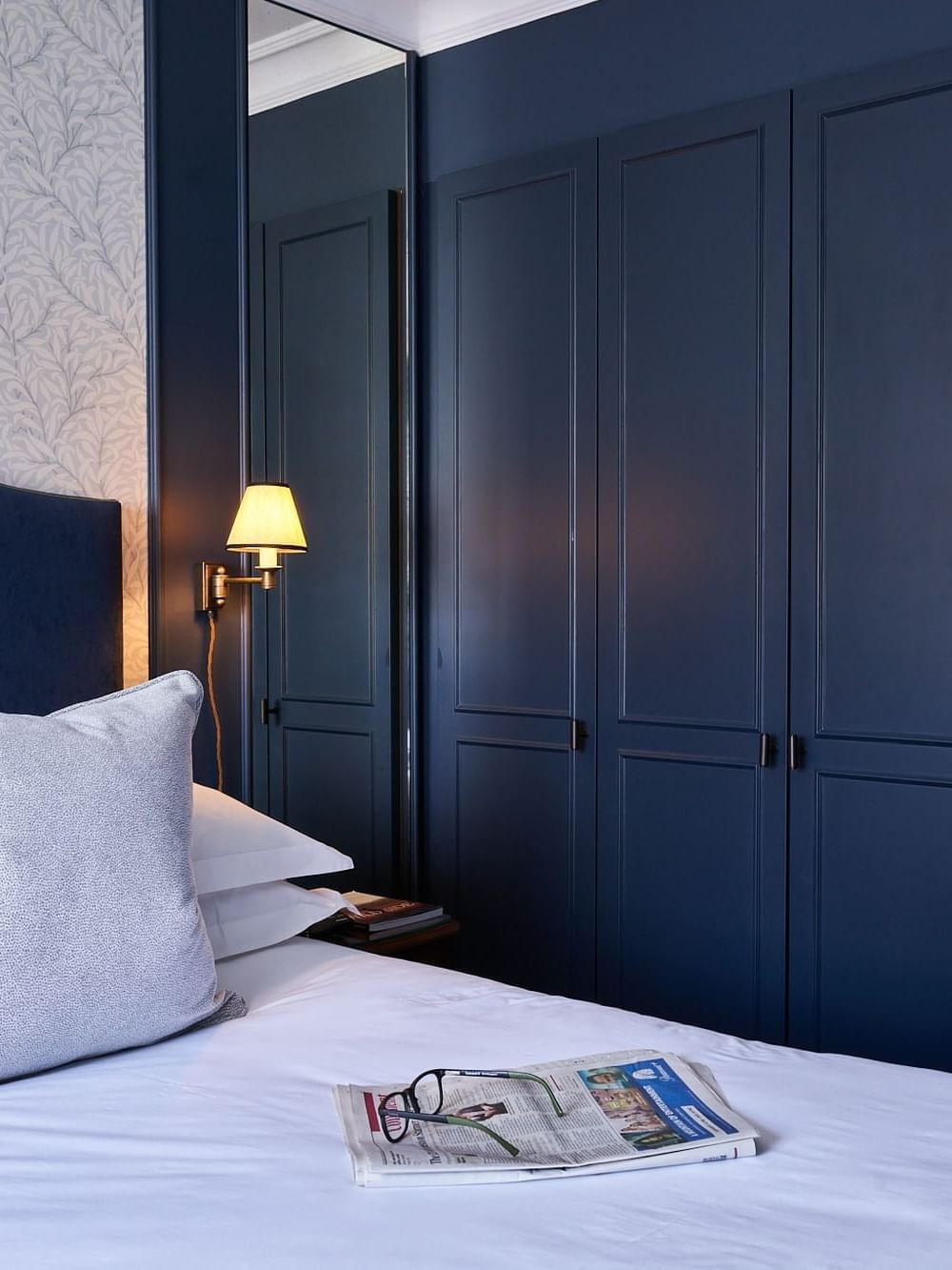 Bedroom with a white bed and glasses on a newspaper by a lamp at The Capital Hotel, Apartments and Townhouse
