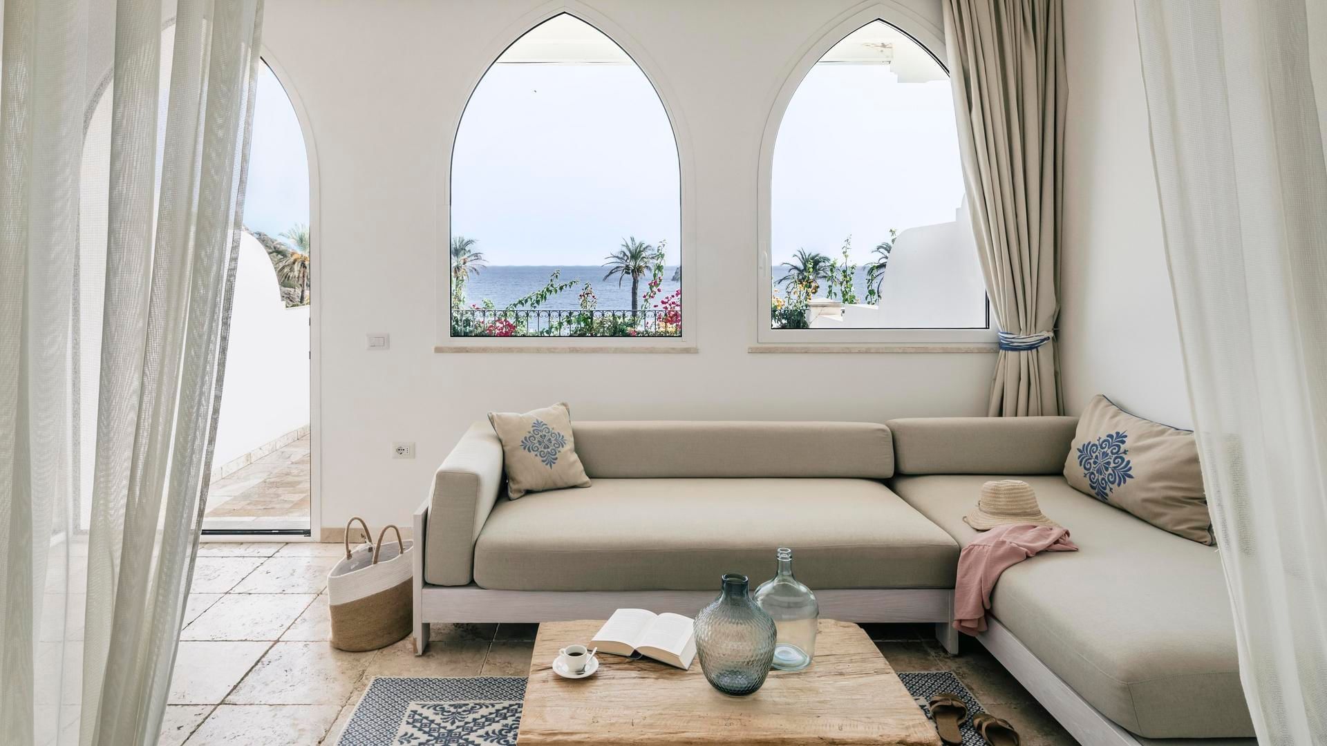 Living room with sofa, sea view through arched windows in Junior Suite with sea view at Falkensteiner Resort Capo Boi