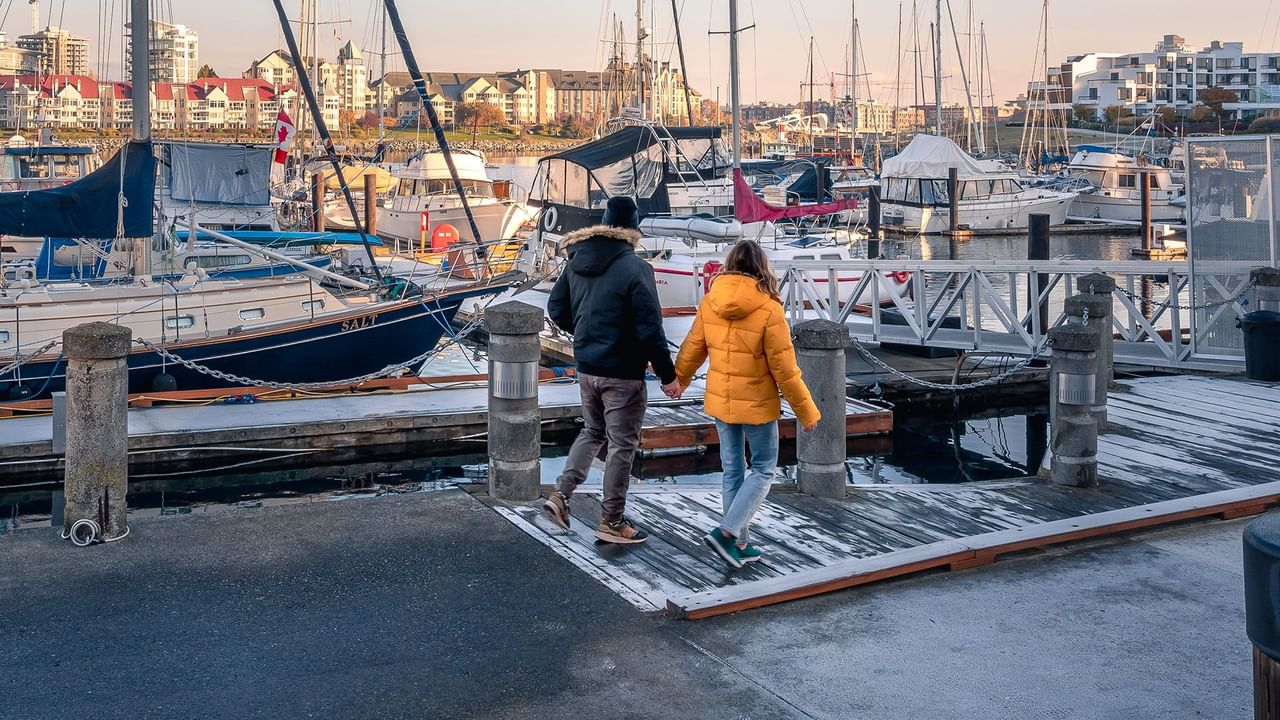 A couple walking along a marina boardwalk past docked sailboats.