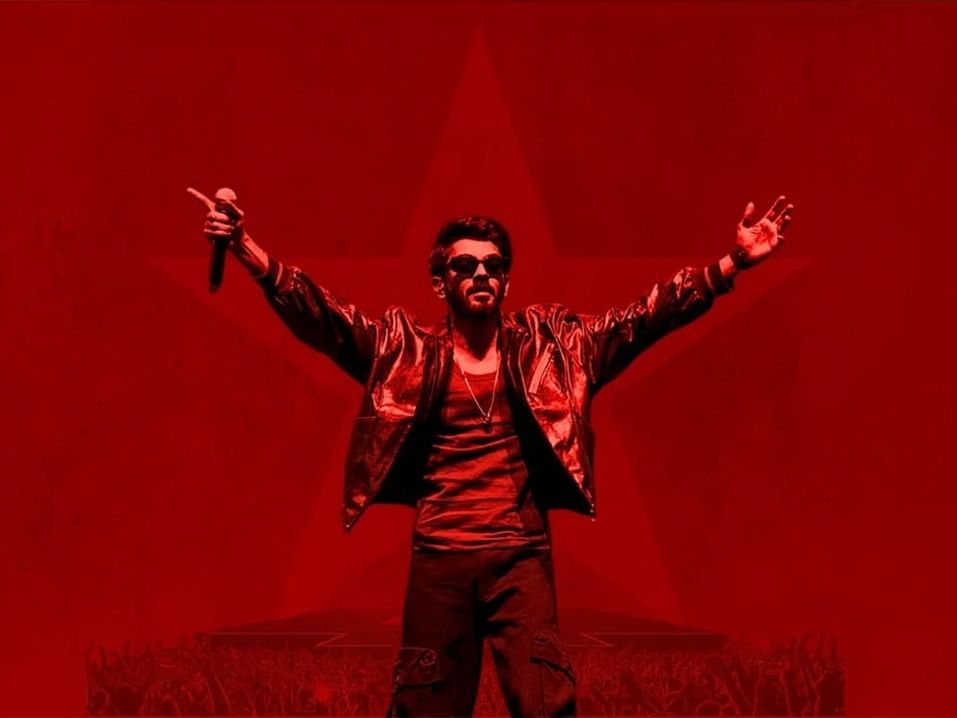 Anirudh performing on stage with raised arms and a microphone against a red background.