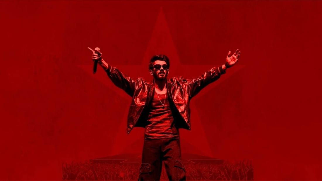 Anirudh performing on stage with raised arms and a microphone against a red background.