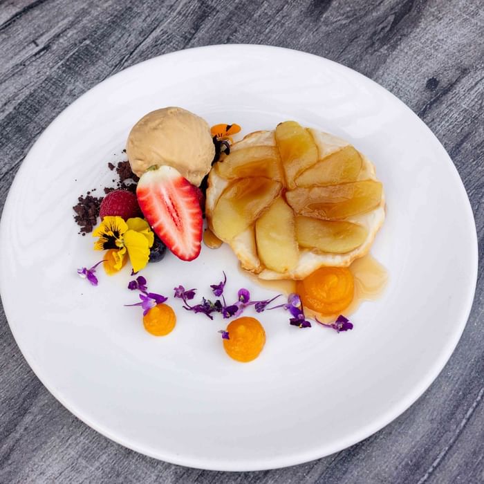 A white plate with a dessert featuring sliced fruit, chocolate, ice cream, and decorative flowers on a wooden table.