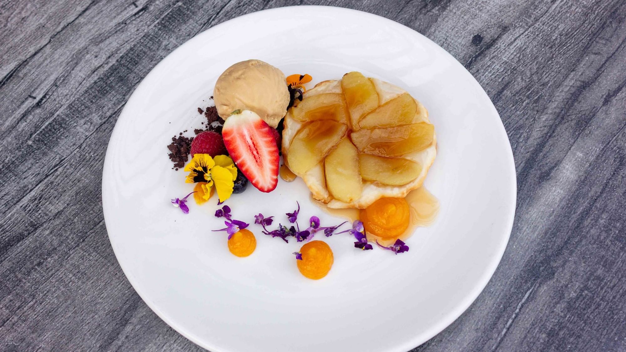 A white plate with a dessert featuring sliced fruit, chocolate, ice cream, and decorative flowers on a wooden table.