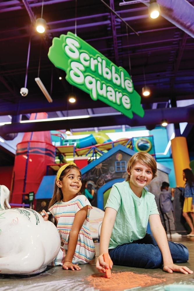 A boy and a girl sitting on the floor of a colorful inside space coloring the floor with chalk under a sign that says Scribble Square.