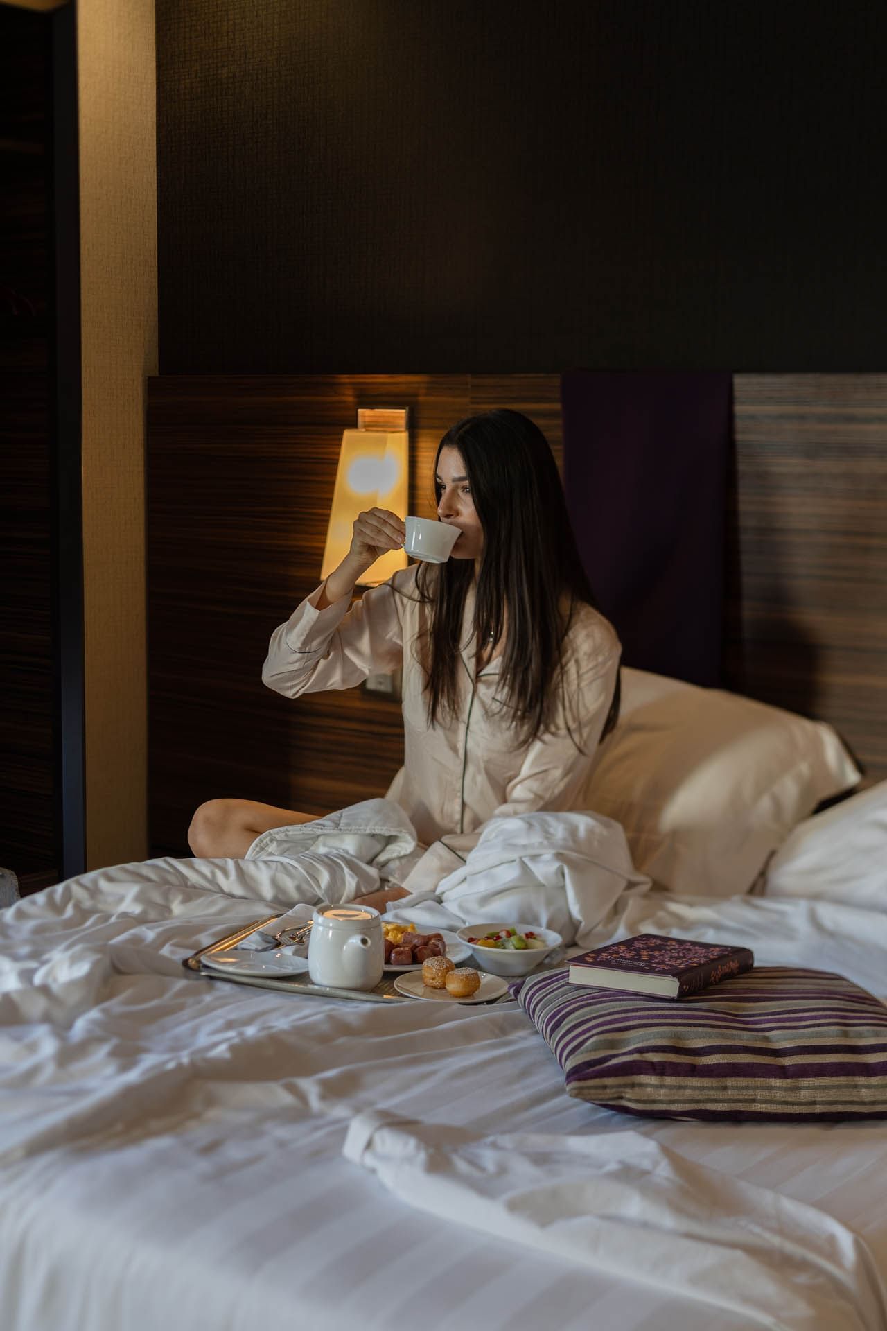 Girl seated in bed wearing pajama shirt enjoys a cup of coffee, surrounded by a tray of breakfast at The Republic Hotel