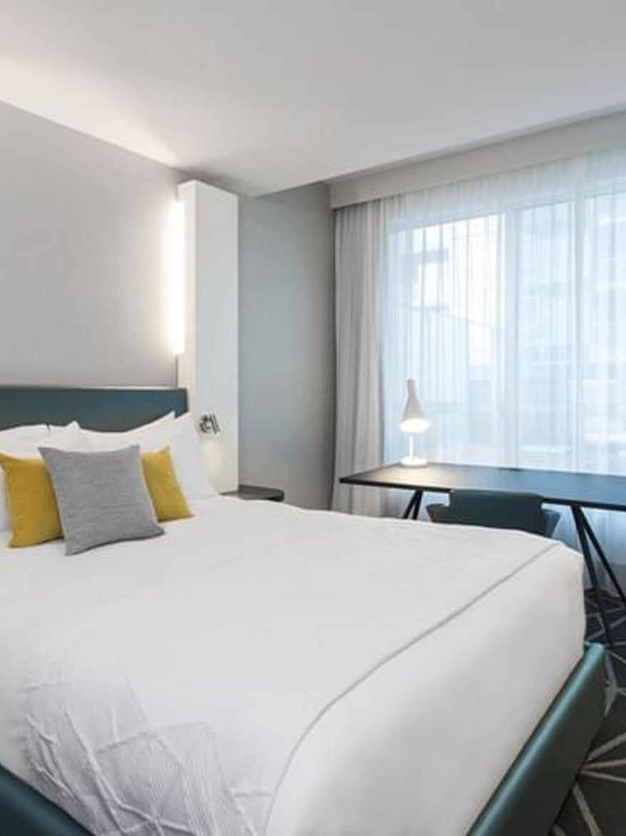 White king-size bed with yellow and gray pillows, desk, and window with curtains at Warwick Le Crystal – Montreal.