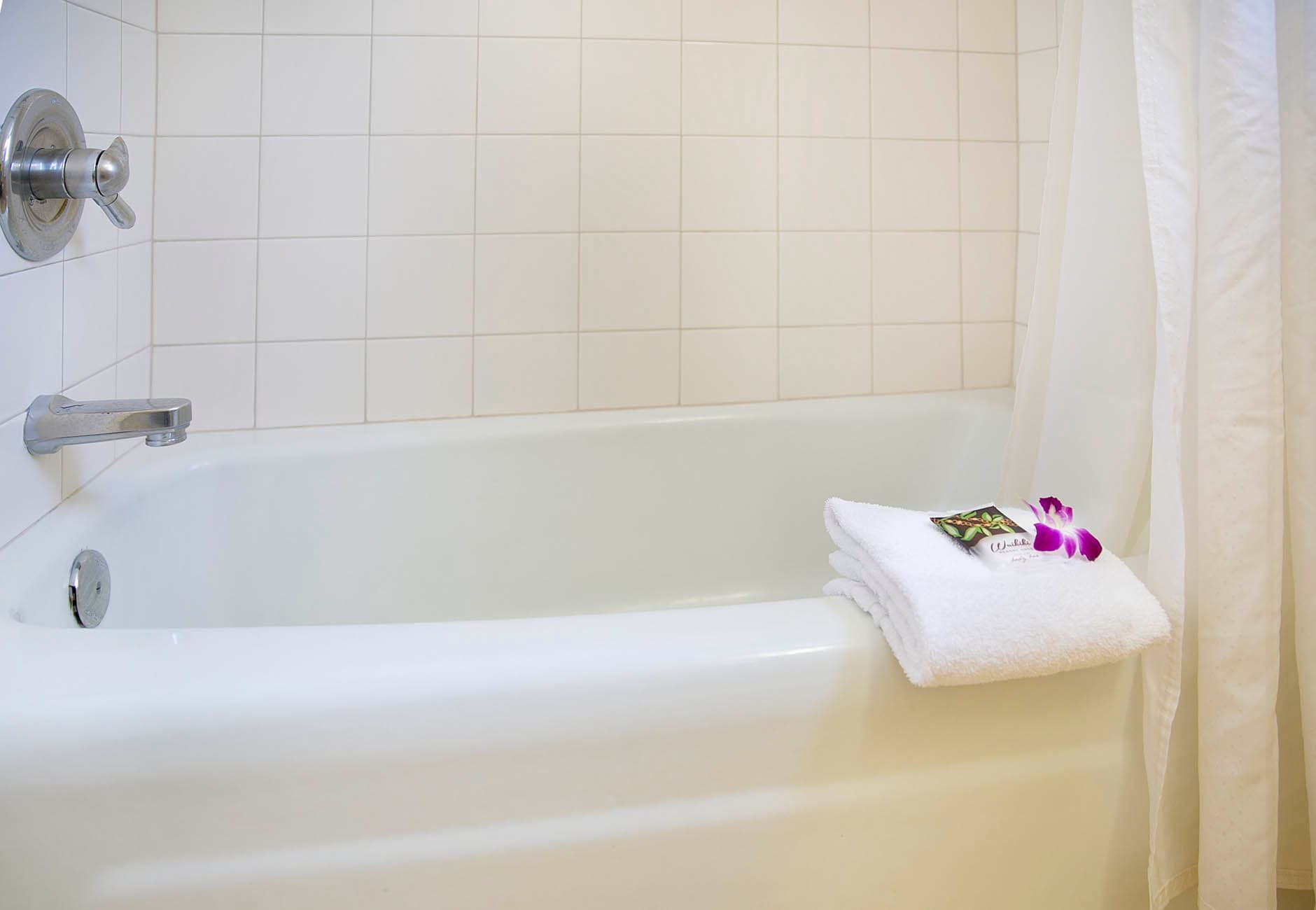 A clean white bathtub with a tiled wall and shower curtain, accompanied by towels and soap.