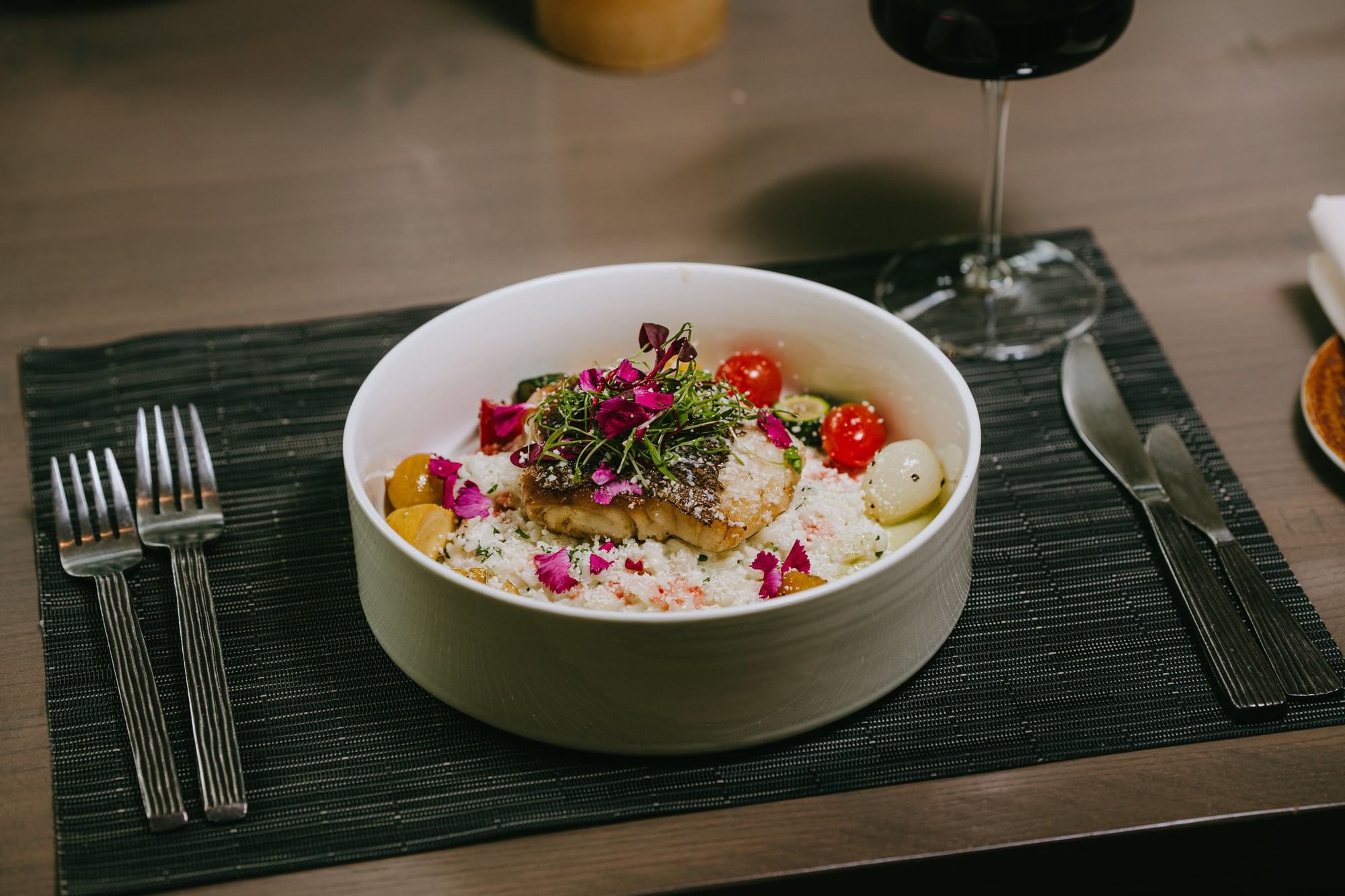 Bowl of seafood salad with rice, garnished with herbs and flowers, accompanied by cutlery and wine glass.