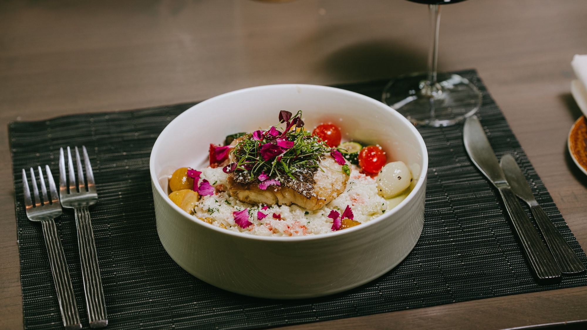 Bowl of seafood salad with rice, garnished with herbs and flowers, accompanied by cutlery and wine glass.