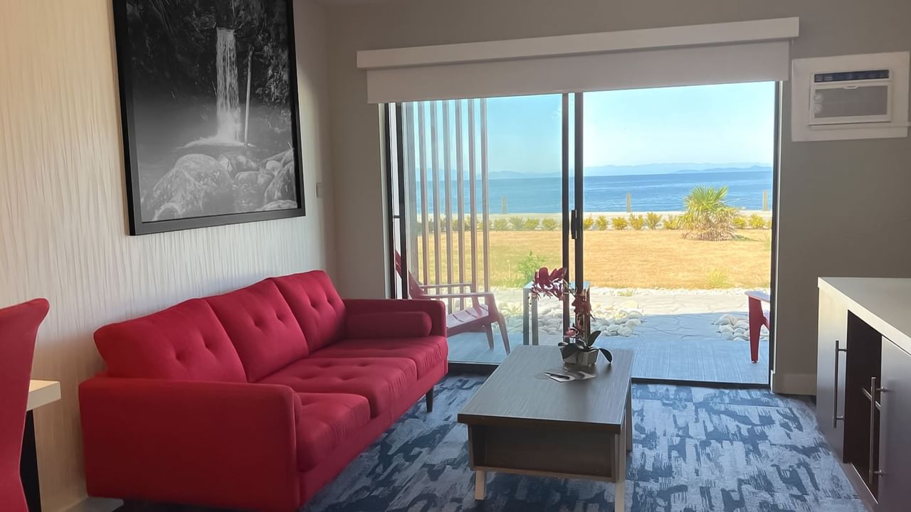 Guest room living area with red couch, coffee table, tv, patio doors, ocean view and patio with red deck chairs