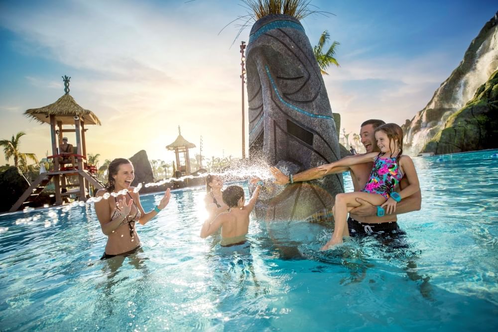 A man, woman, and three children play in a pool with a tiki-inspired fountain shooting water beside them. 