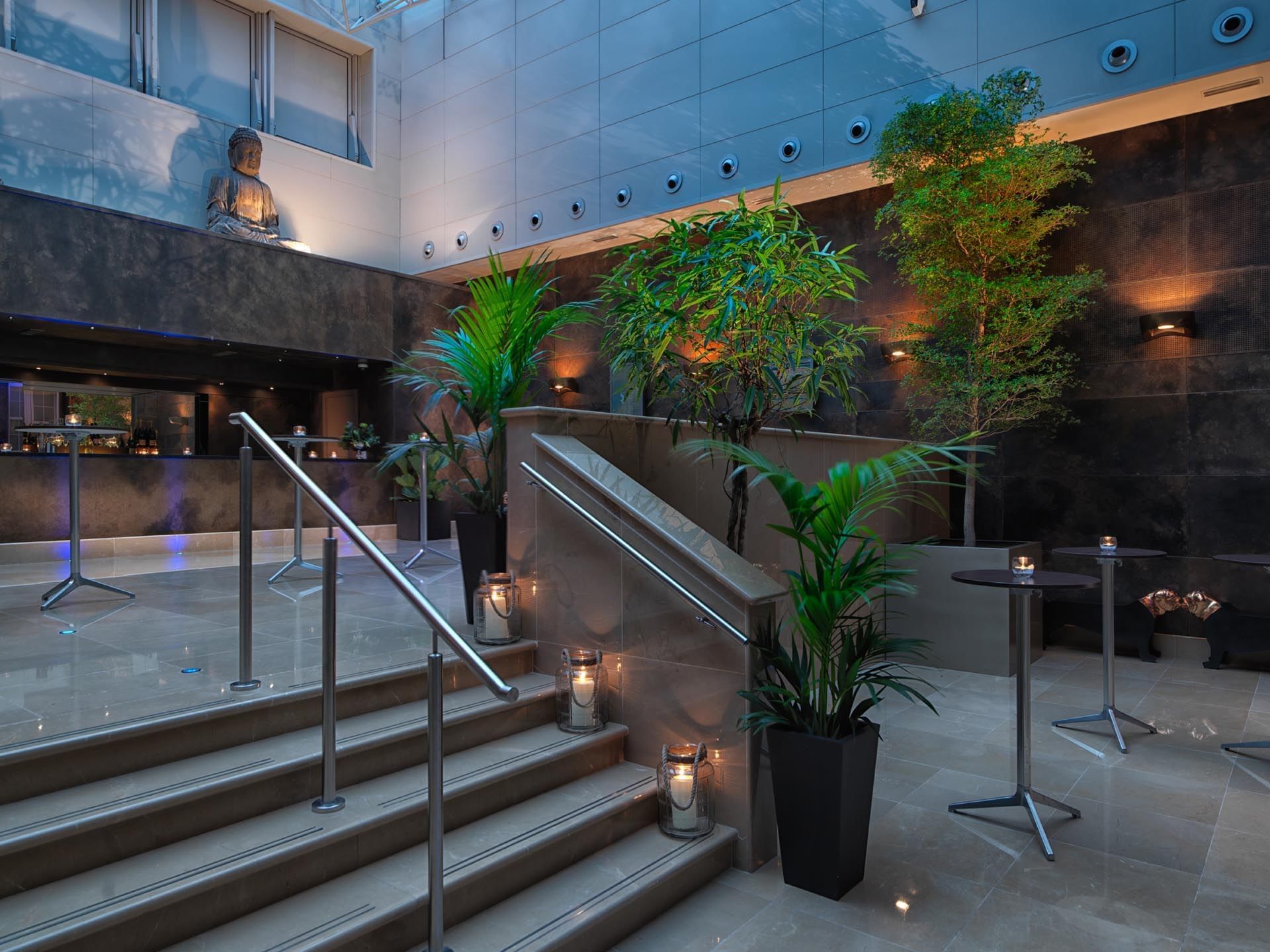 The May Fair Conference Atrium with a staircase, ambient lighting, and lush potted plants at The May Fair Hotel