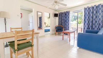 One-bedroom living room with blue sofa beside wooden chairs & patterned curtains near patio doors at Worthing Court Apartment
