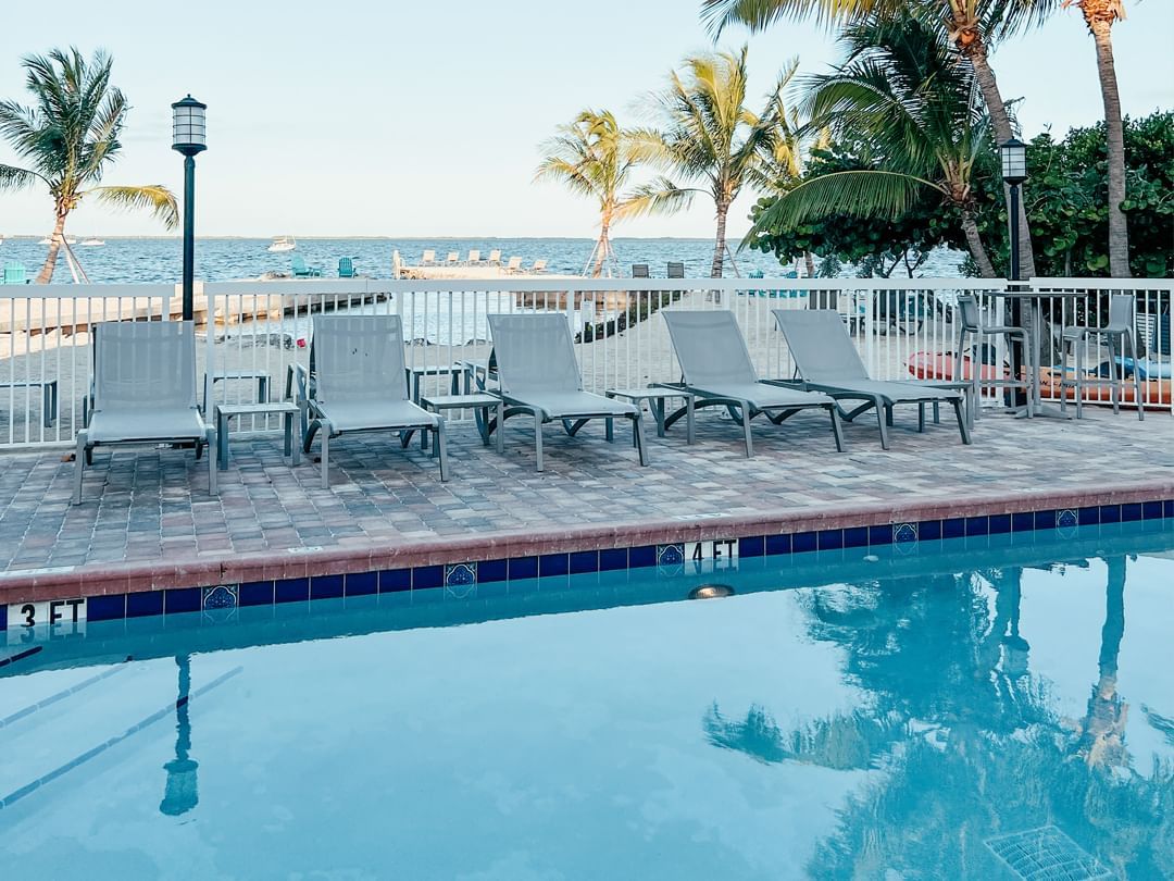 Poolside lounge chairs and tables next to a fence overlooking a beach and ocean.