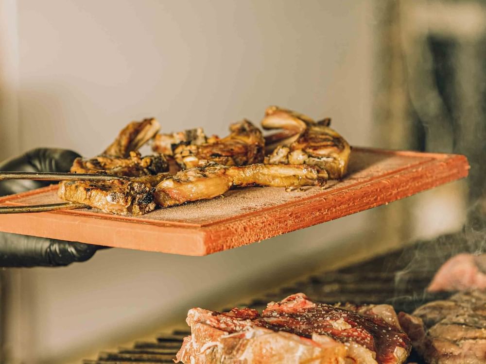 A person with black gloves holding a tong and a wooden board with meat above grill.