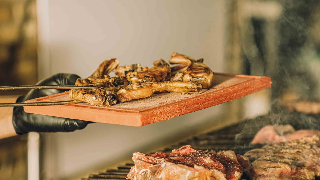 A person with black gloves holding a tong and a wooden board with meat above grill.