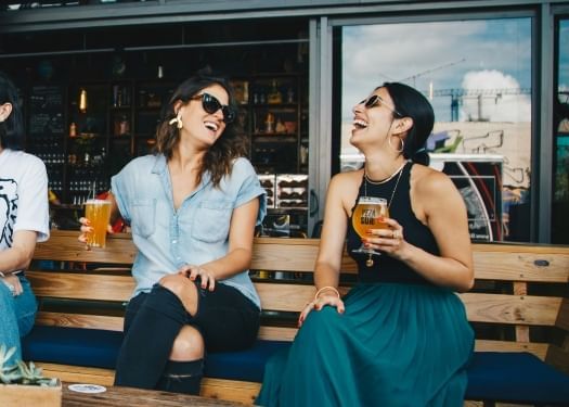 Two women holding beers in their hands, laughing and sitting on a bench at an outdoor bar. 