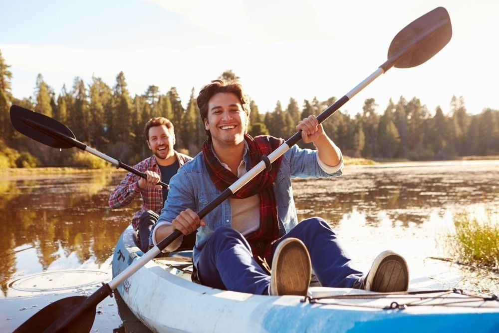 Two handsome men sit in a kayak on a lake, their oars mid-paddle.