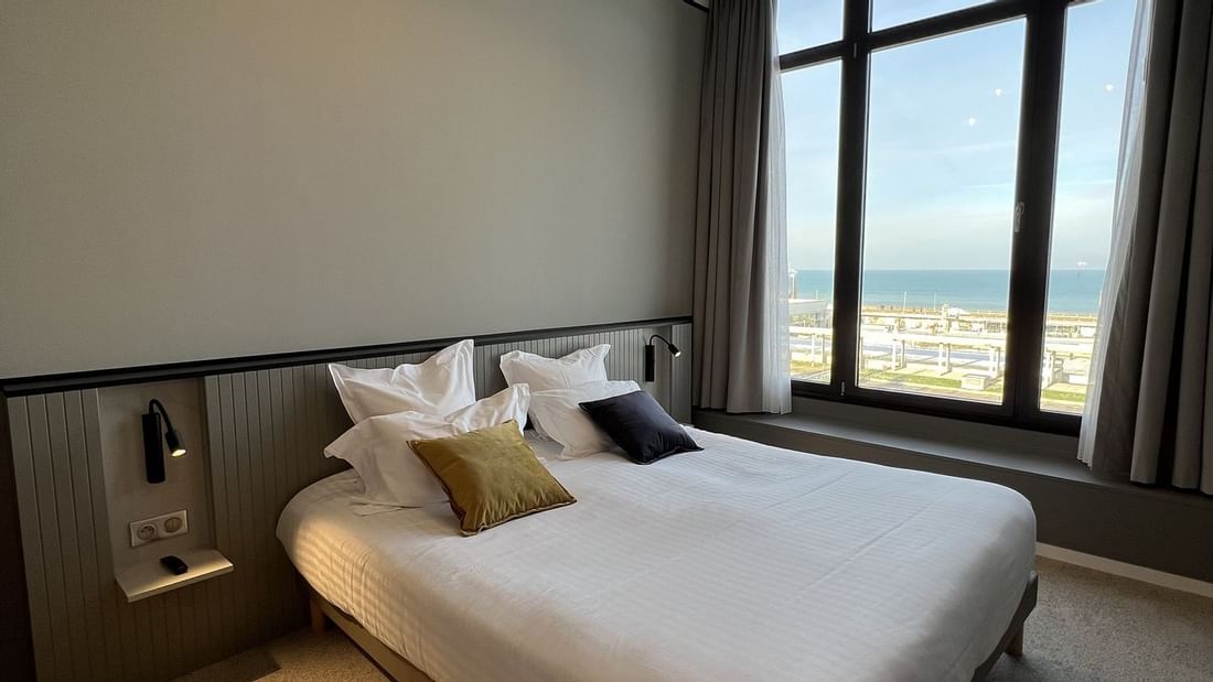 Chambre avec lit, coussins, fenêtre vue mer, lampe et moquette au sol.
