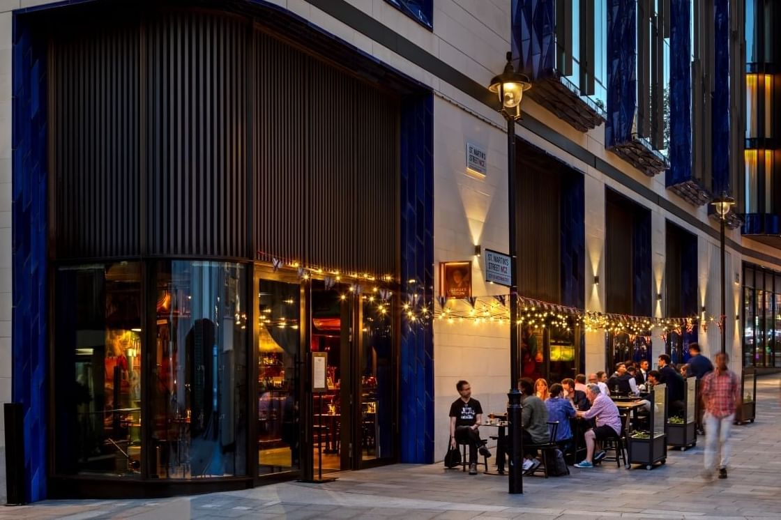 Patrons dining at outdoor tables under string lights on a street corner at dusk outside of The Londoner Hotel