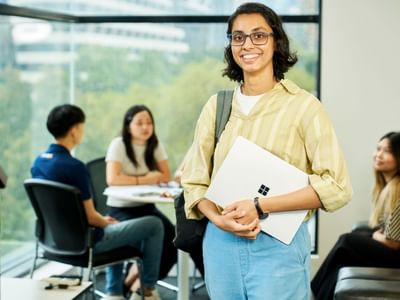 A resident holding a laptop stands smiling with others at tables behind her.