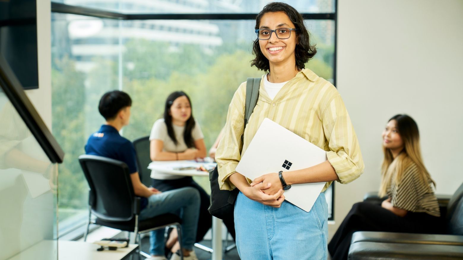 A resident holding a laptop stands smiling with others at tables behind her.