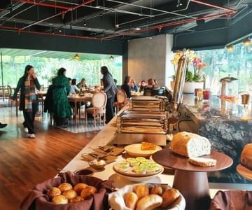 Breakfast buffet station with fresh bread and pastries near guests dining by a scenic window inside Hotel B3 Sibelius