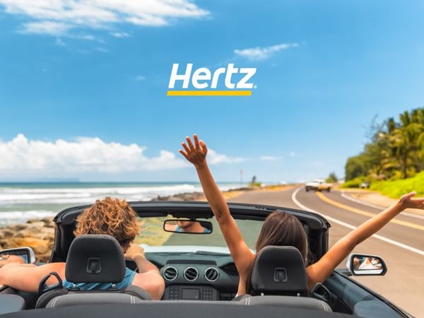 Banner de Hertz con pareja conduciendo un convertible en carretera costera cerca de Fiesta Inn, levantando los brazos