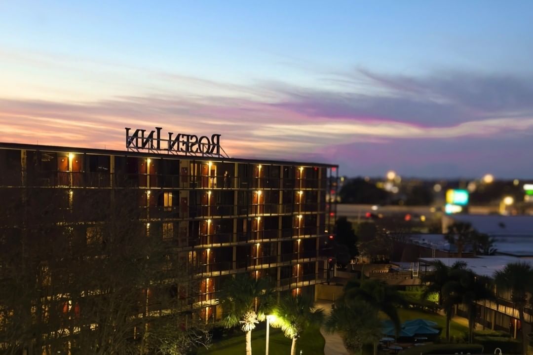 A glowing hotel building at twilight with a view of the back of a large sign atop the roof reading Rosen Inn.