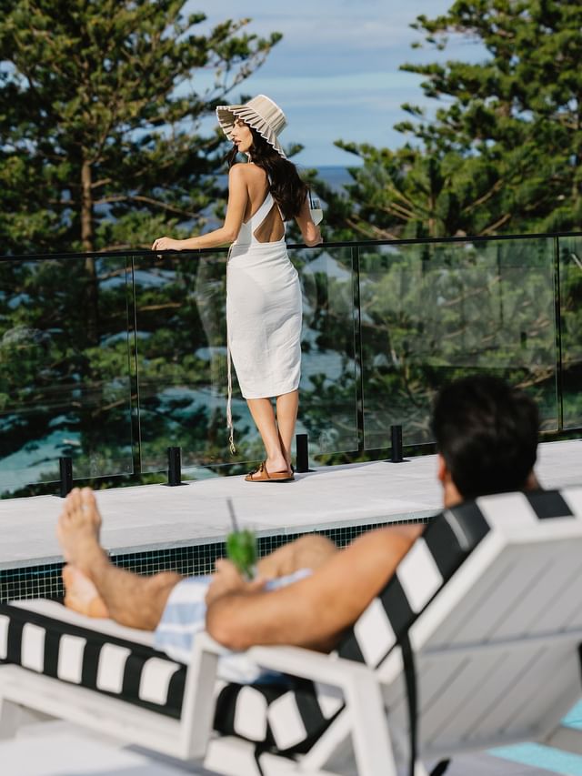 Woman at Elysium Manly standing in a white wrap by a glass pool fence to overlook pine trees, a top-choice hotel in Manly