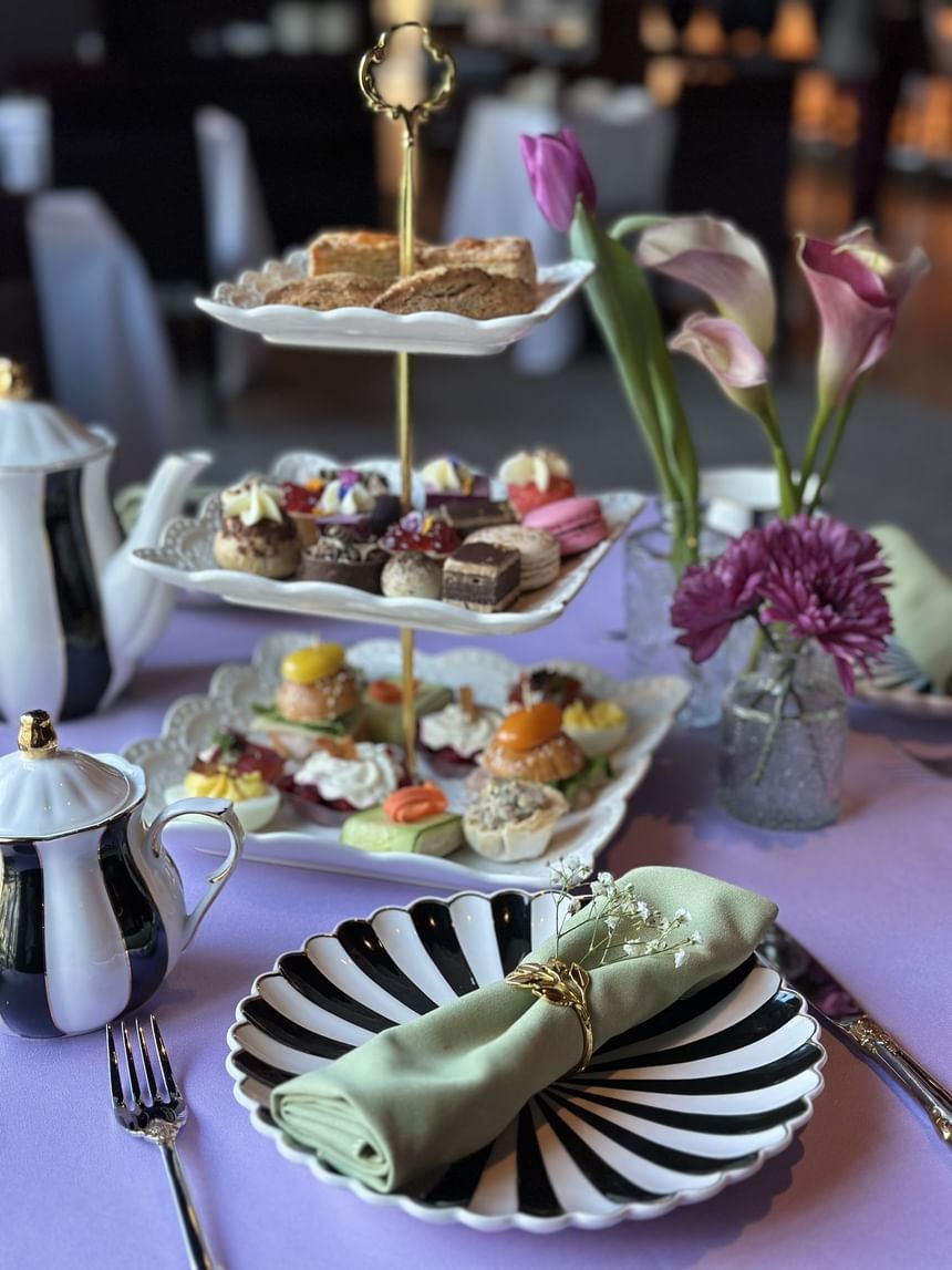 A plate with a napkin, a teapot, a vase with flowers, and a three-tiered tray with desserts.