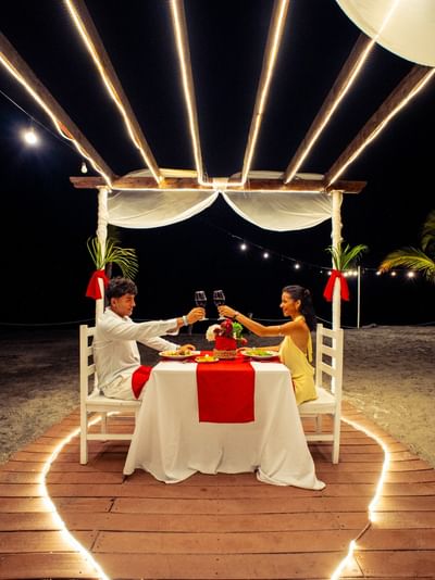 Couple toasting at illuminated beachfront dinner table under wooden pergola at night at Playa Blanca Beach Resort