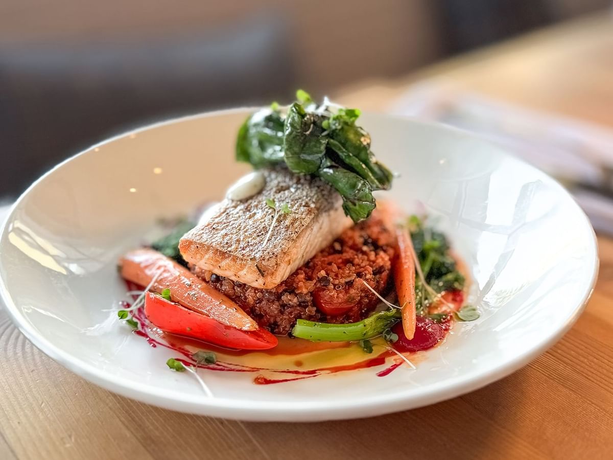A plate with a fish, vegetables, and greens on a wooden table.