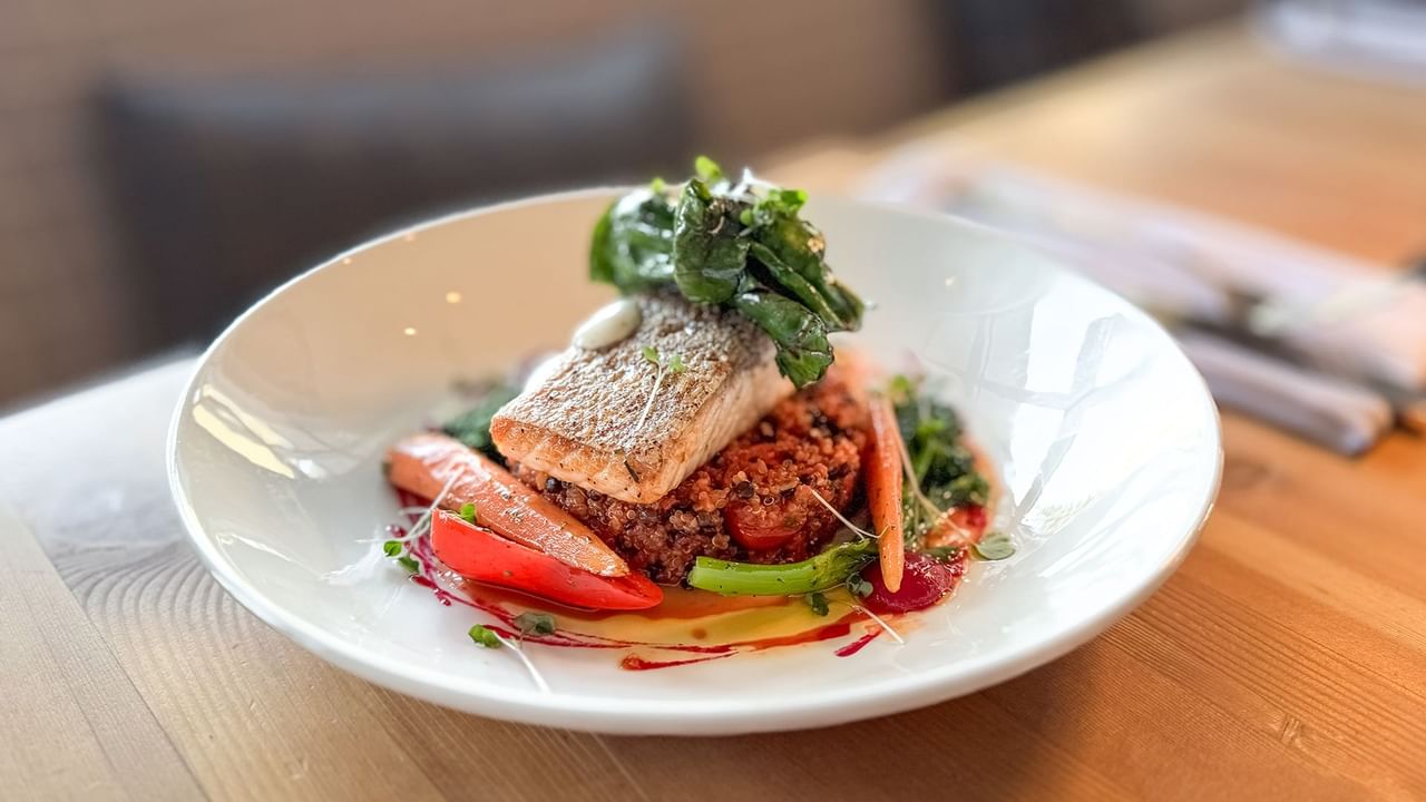 A plate with a fish, vegetables, and greens on a wooden table.